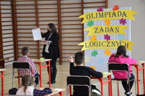 Nauczyciel pokazuje dzieciom karty zadań, w tle tablica z napisem: Olimpiada zadań logicznych